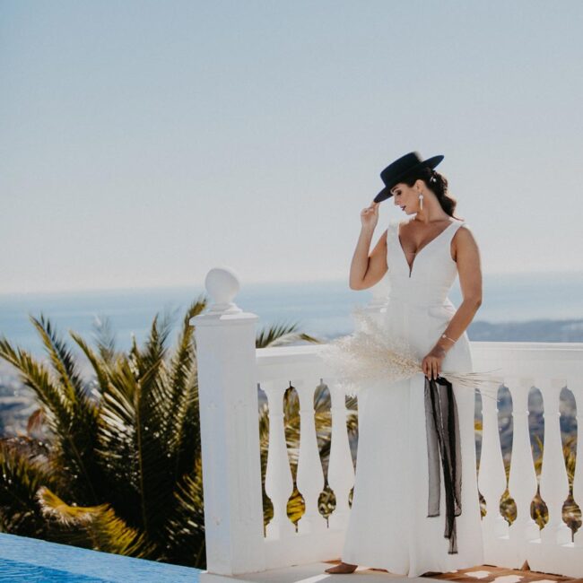 Destination Wedding Shooting Hochzeit in Spanien mit Urlaubsfeeling. Braut in einem modernen Hochzeitskleid und schwarzen Hut steht neben dem Pool