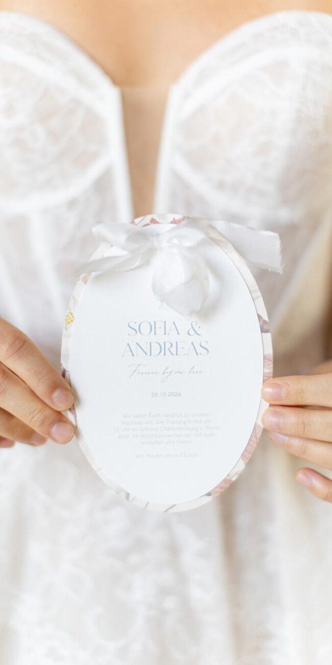 Braut hält eine ovale Hochzeitseinladung mit Seidenband vor ihrem Brautkleid
