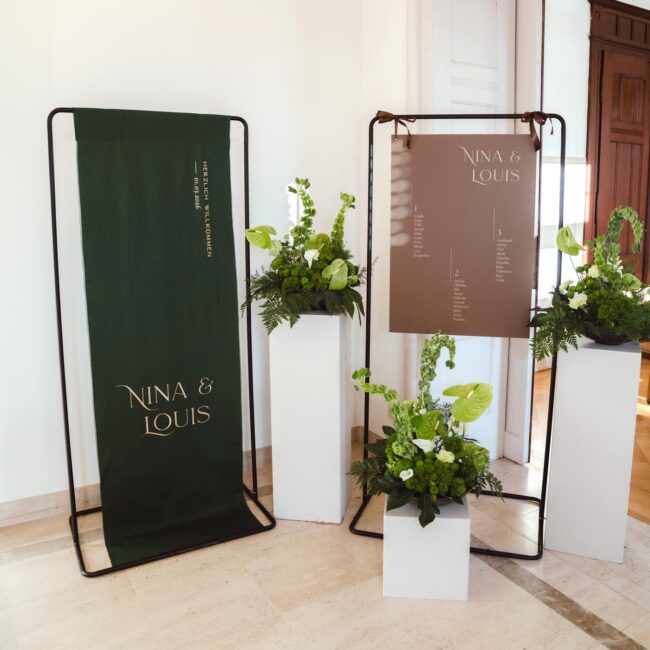 Individuelle Hochzeitsschilder bestehend aus einem Stoffbanner in grün und goldener Schrift als Willkommensschild und ein Sitzplan Schild in Hellbraun